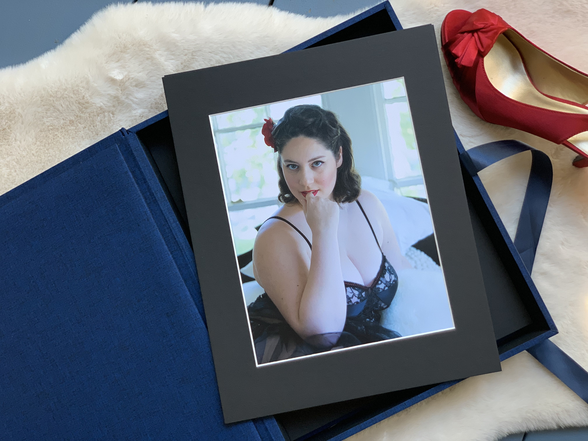 A framed portrait of a woman in lingerie with a red flower in her hair is placed in an open blue box next to a red high-heeled shoe on a fur surface. House of Secrets Boudoir Photography by Tanya Eivin