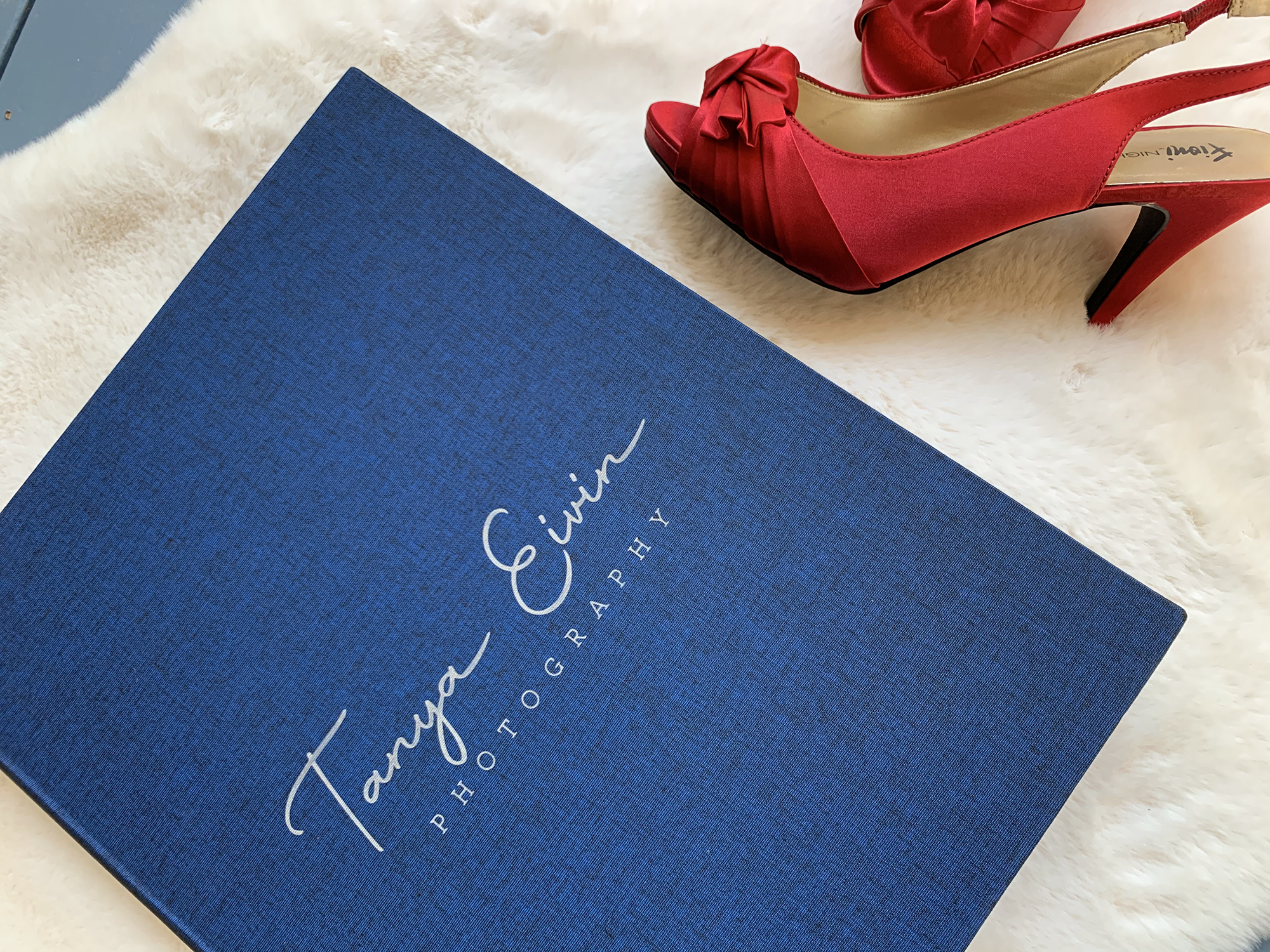 A blue photography portfolio book labeled "Tanya Eivin Photography" lies on a white fur surface next to a pair of red high-heeled shoes. House of Secrets Boudoir Photography by Tanya Eivin