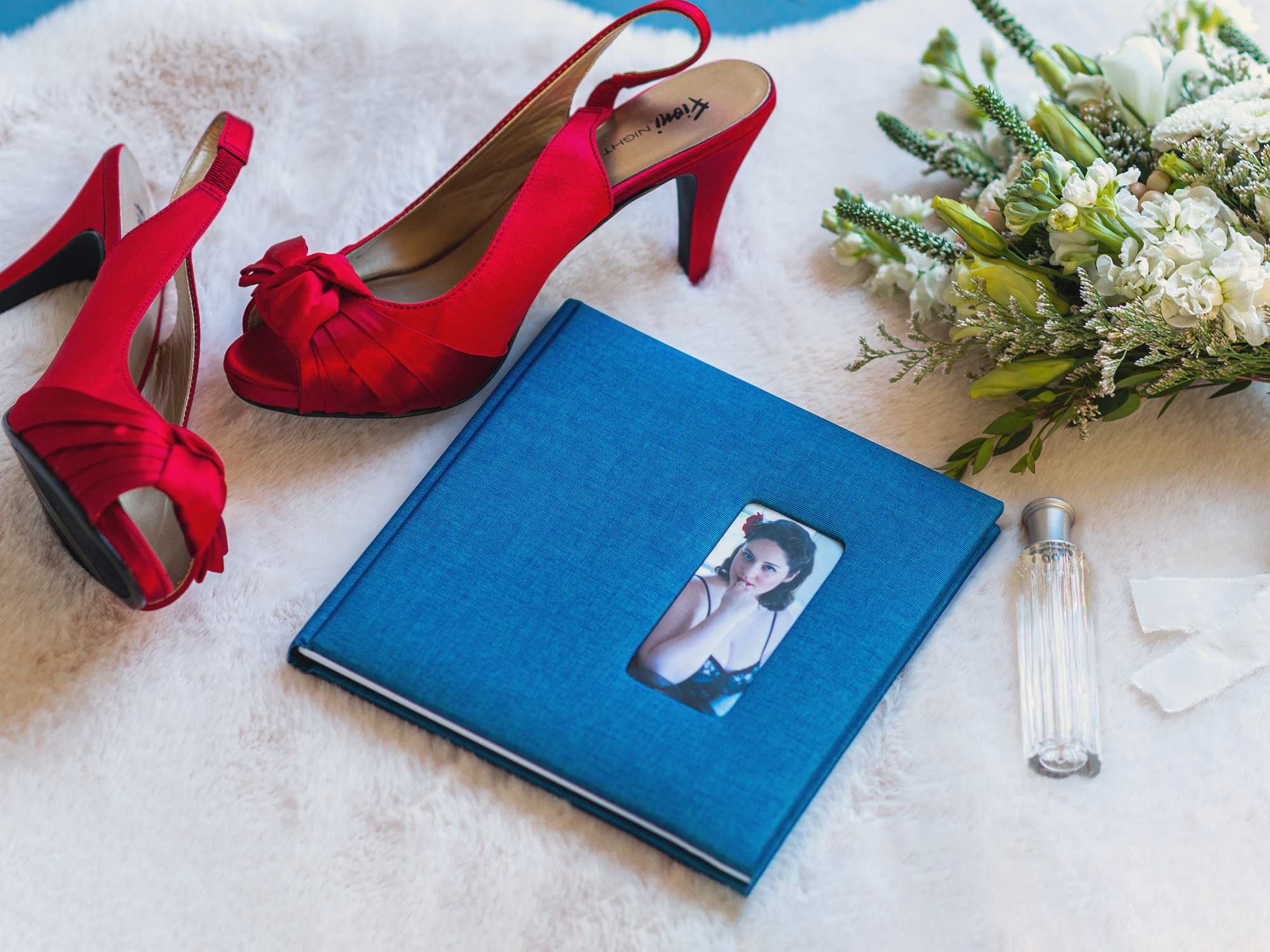 A pair of red high heels, a blue photo album with a woman's portrait, a bouquet of white flowers, and a perfume bottle arranged on a white furry surface. House of Secrets Boudoir Photography by Tanya Eivin