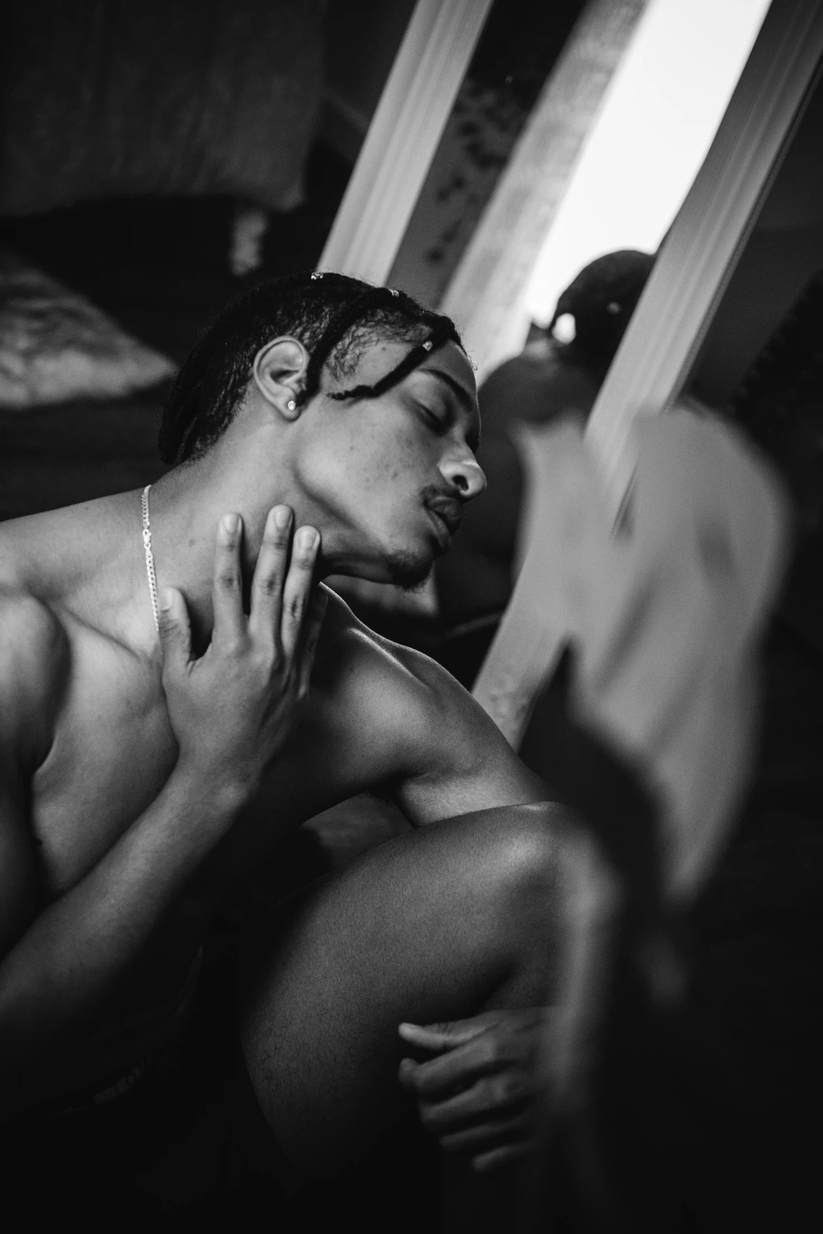 Black and white photo of a shirtless person sitting on the floor, touching their neck, with a mirror and blurred foreground visible.