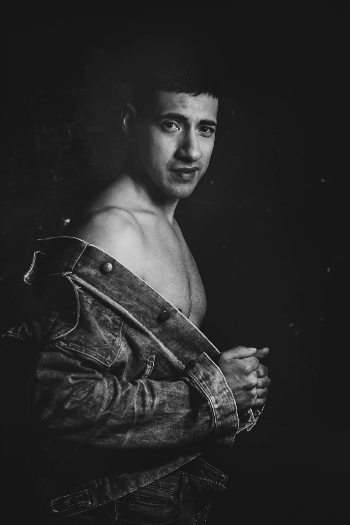 A man poses against a dark background, wearing a denim jacket off his shoulders, looking at the camera with a neutral expression.