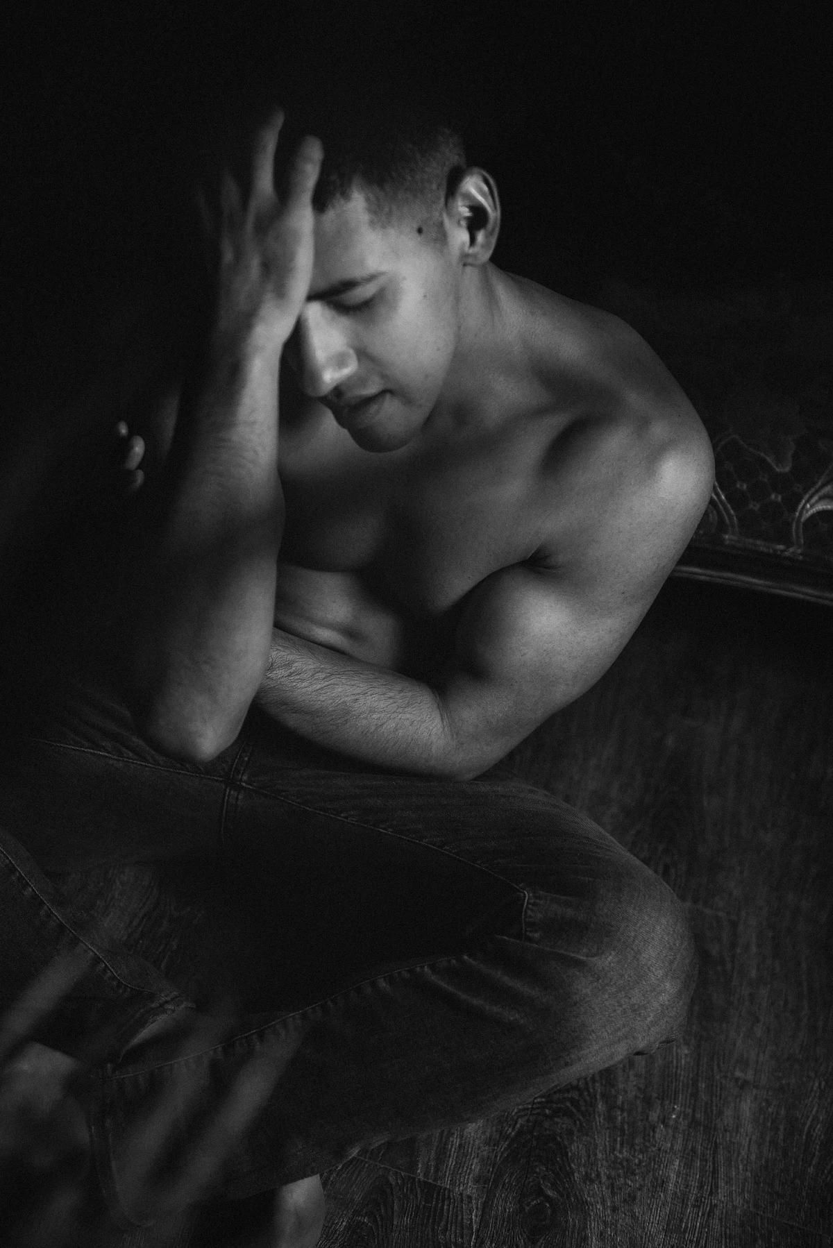 A shirtless man sits on the floor in low lighting with his head down, one hand on his forehead, and his other arm across his body.
