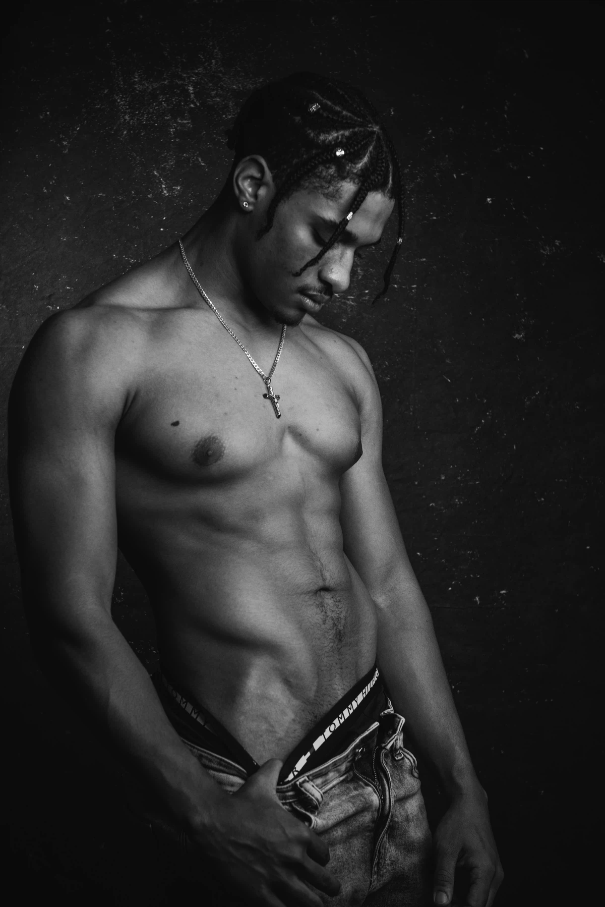 Shirtless man with a necklace poses against a dark background, looking down with one hand resting on his jeans.