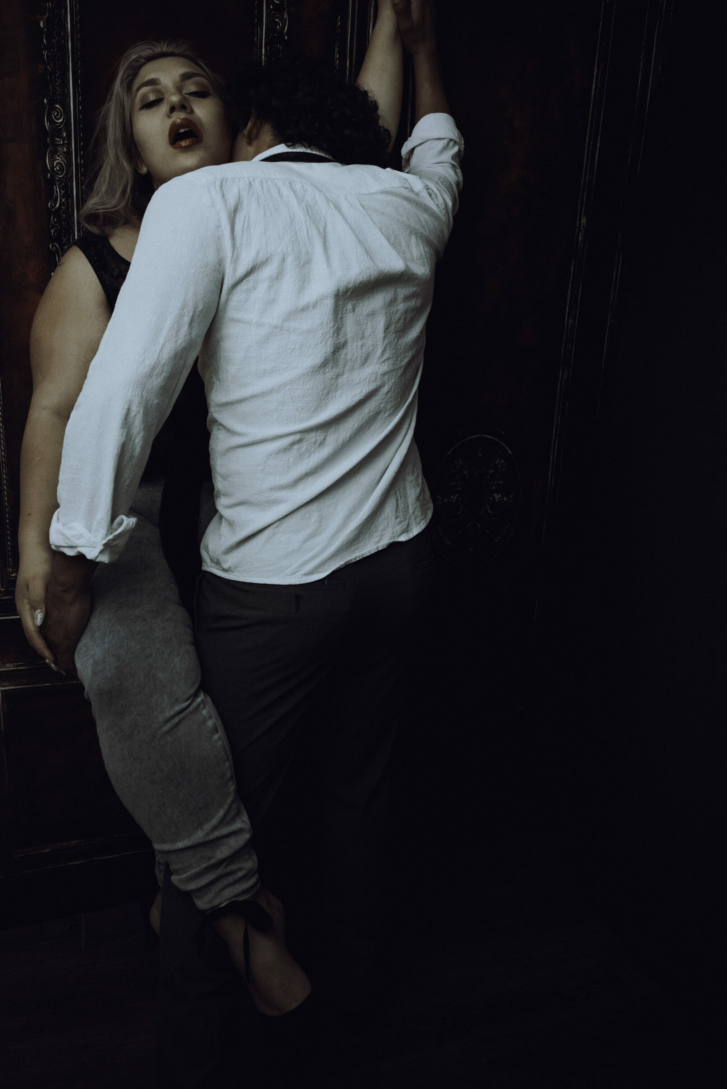 A woman leans against a dark wall with her arm raised as a man in a white shirt and gray pants stands close to her