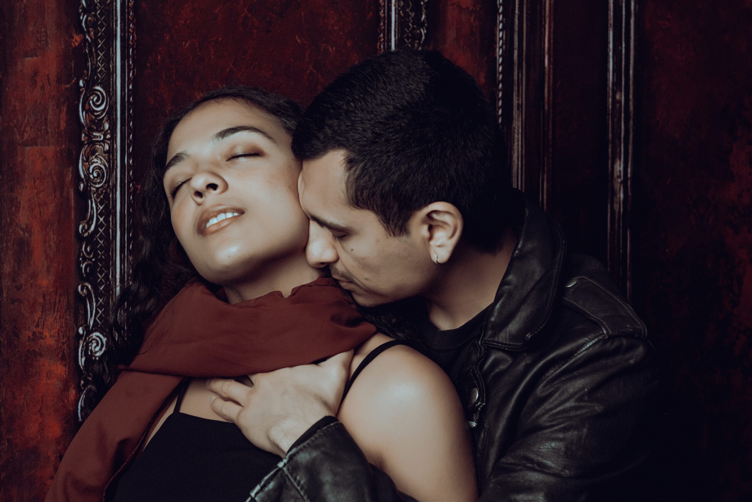 A man in a black leather jacket embraces a woman with closed eyes and a red scarf, standing against an ornate wooden background.