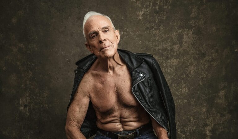 Older man with gray hair sits barefoot on a box wearing jeans and an open leather jacket, chest exposed