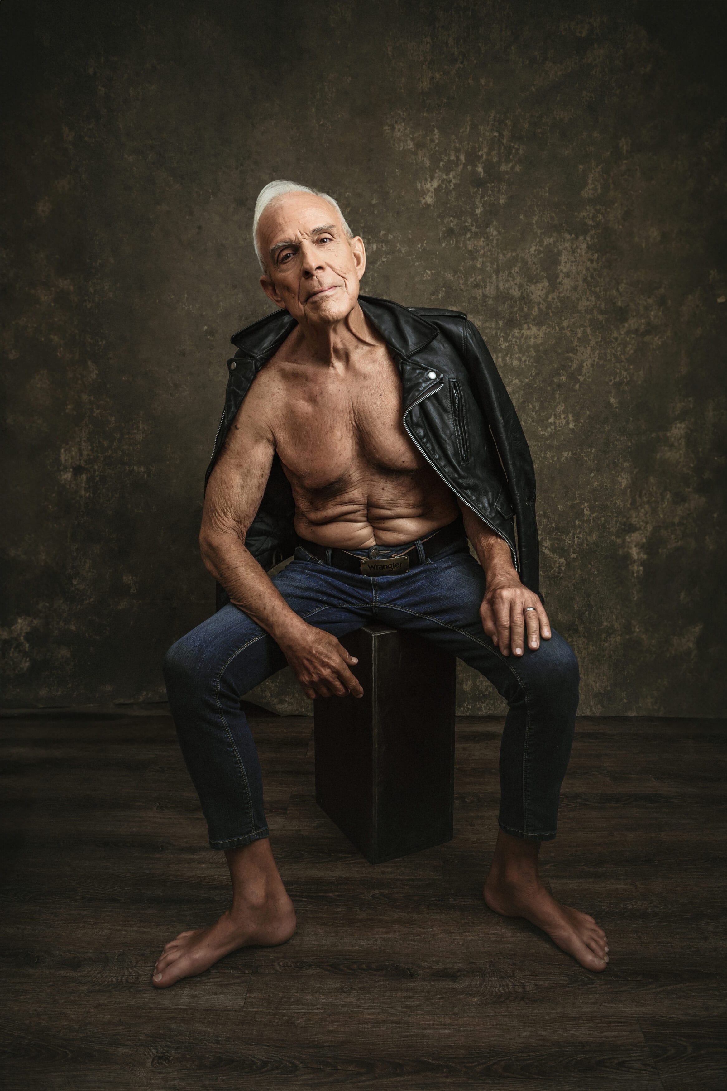 Older man with gray hair sits barefoot on a box wearing jeans and an open leather jacket, exposing his chest, against a textured brown studio backdrop. house of secrets boudoir photography mens boudoir photography