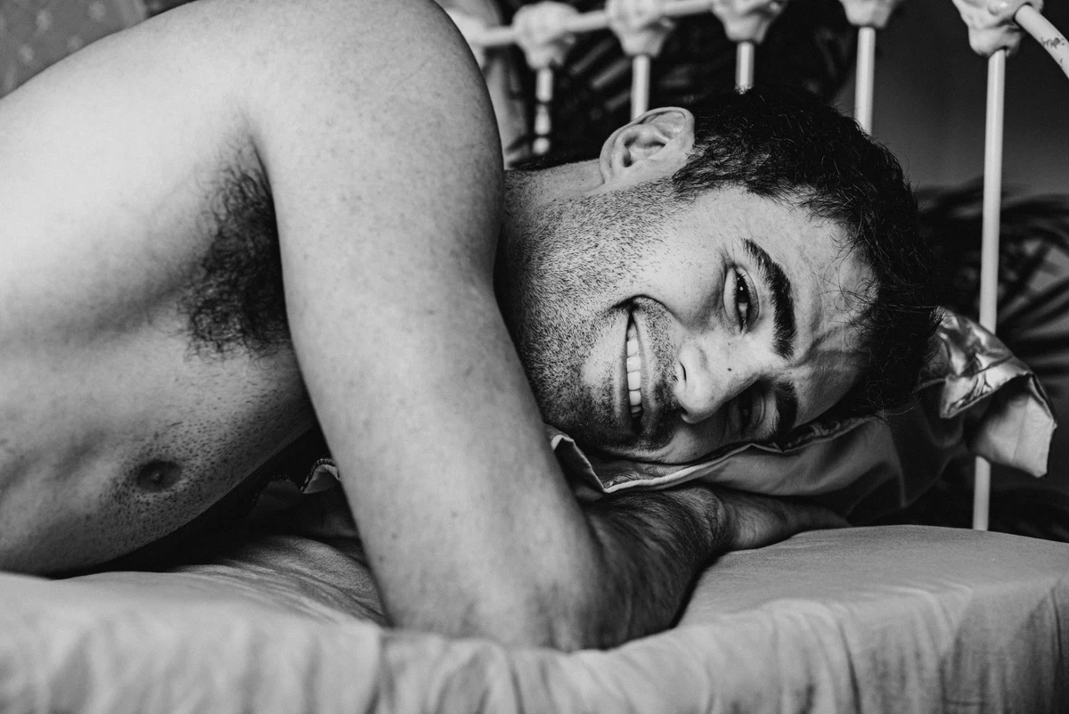 A shirtless man lies on a bed, resting his head on a pillow and smiling, in a black and white photo.