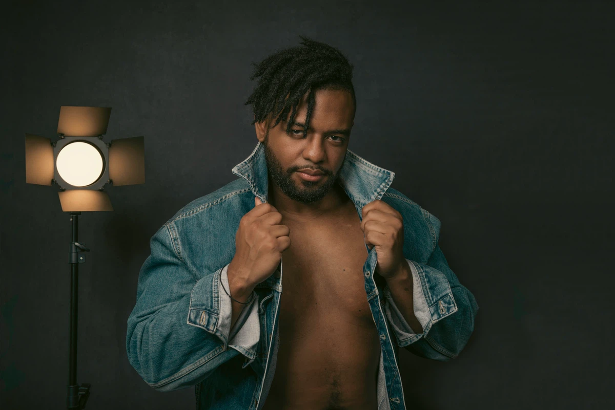 Man poses in front of a dark background, wearing an open denim jacket and holding its collar