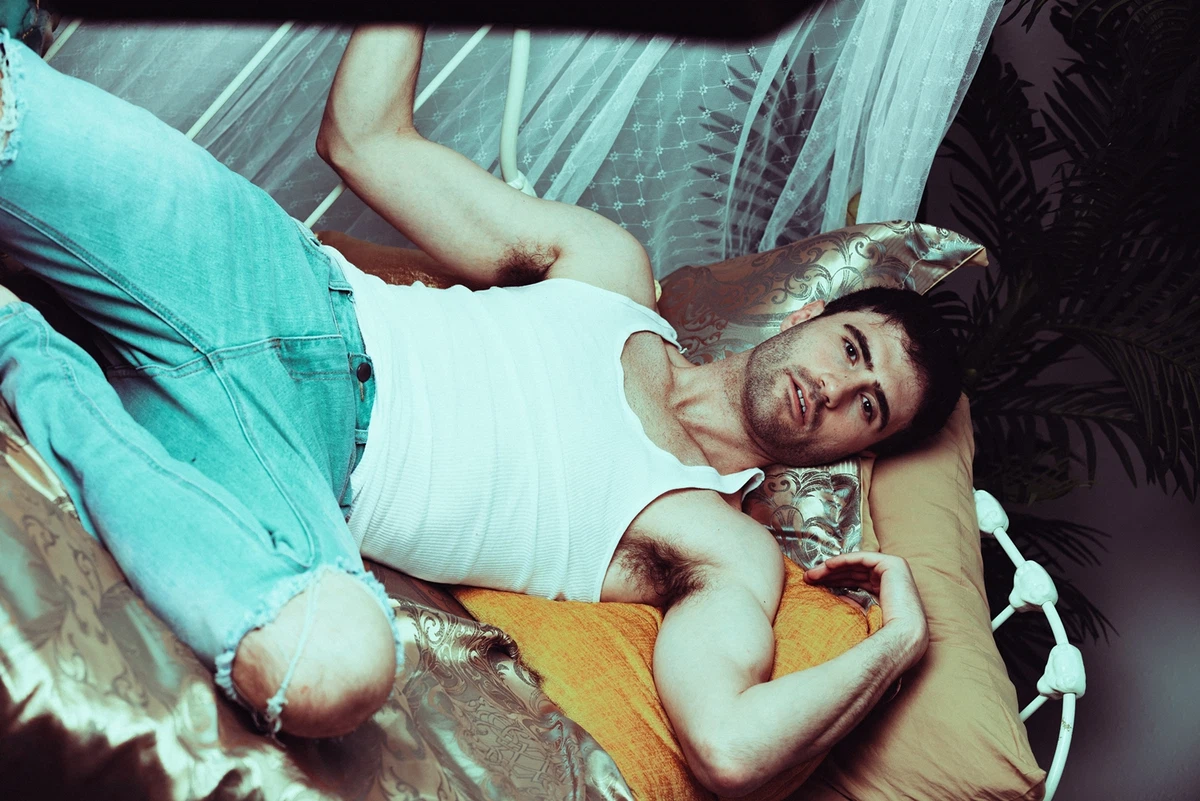 A man in a white tank top and ripped jeans lies on a bed with gold bedding and sheer curtains, looking at the camera.