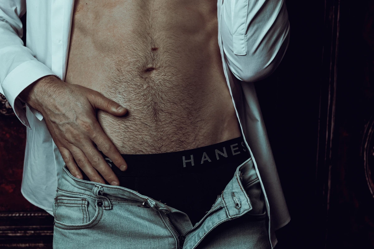 A man wearing a white shirt and partially unzipped jeans shows the waistband of black Hanes underwear.