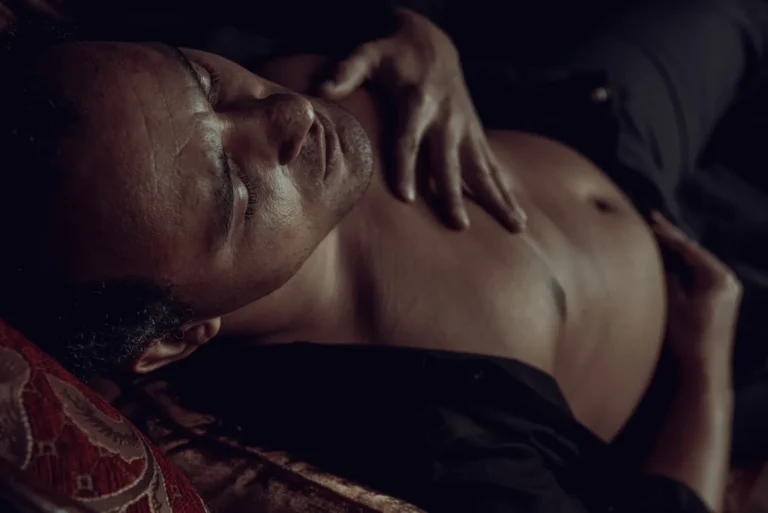 A man with closed eyes lies on a settee, shirt partially open, with one hand on his chest and the other on his abdomen.