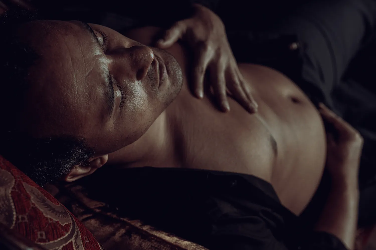 A man with closed eyes lies on a settee, shirt partially open, with one hand on his chest and the other on his abdomen.