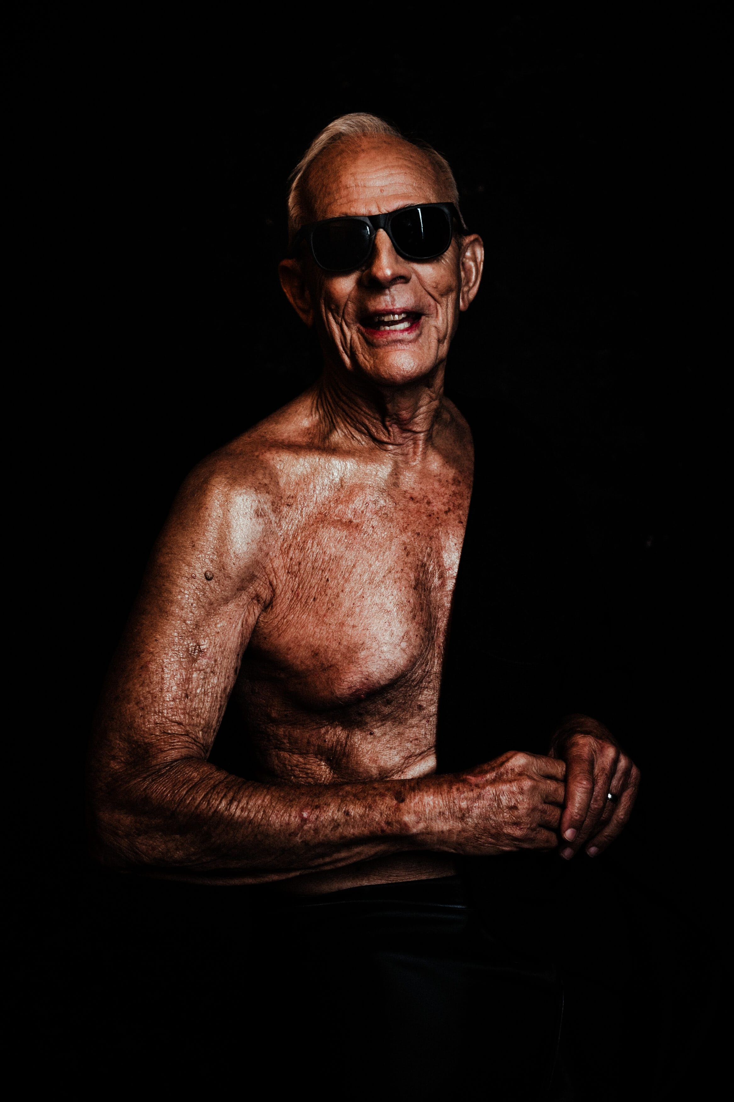 Older man with short white hair, bare chested, wearing dark sunglasses and a black cloth over one shoulder, smiling against a dark background. house of secrets boudoir photography showing mens boudoir photography